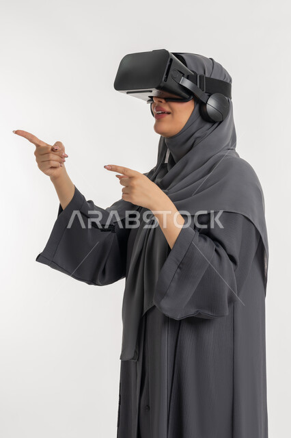 محاولة النقر على شيء ما, المتعة  بالمشاهدة عبر نظارة الواقع الافتراضي, بورتريه لإمرأة عربية خليجية إماراتية محجبة ترتدي العباءة تضع نظارات ثلاثية الأبعاد, الاندماج ومحاكاة العالم الخيالي, شابة سعودية ترفع اصبع السبابة وتقوم بالاشارة الى مكان معين، خلفية بيضاء