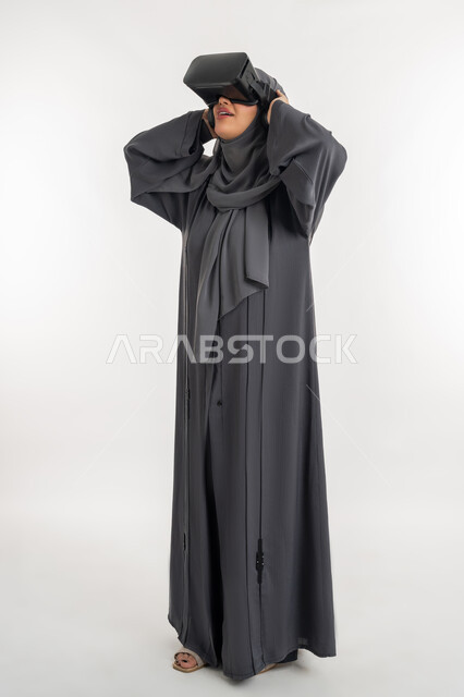 استخدام التكنولوجيا والتقنيات الحديثة، محاكاة العالم الخيالي، شابة سعودية ترفع يديها بايماءات الدهشة والاستغراب، بورتريه لإمرأة عربية خليجية إماراتية محجبة ترتدي العباءة وتضع نظارة الواقع الافتراضي، التفاعل مع فيديو ثلاثي الأبعاد، الطول الكامل للجسم، خلفيه بيضاء