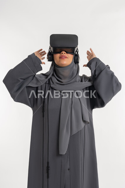 محاكاة العالم الخيالي، مشاهدة الأفلام عبر نظارات في آر، بورتريه لإمرأة عربية خليجية إماراتية محجبة ترتدي عباءة وتضع نظارة الواقع الافتراضي، شابة سعودية ترفع يديها بايماءات الدهشة والاستغراب, التفاعل مع فيديو ثلاثي الأبعاد، استخدام التكنولوجيا الحديثة، خلفيه بيضاء