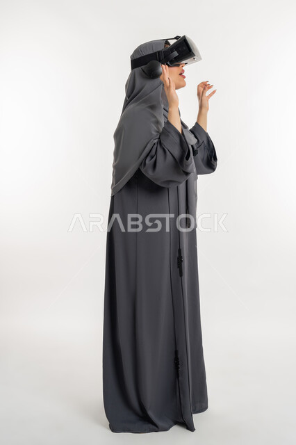 محاكاة العالم الخيالي، شابة سعودية ترفع يديها بايماءات الدهشة والاستغراب، بورتريه لإمرأة عربية خليجية إماراتية محجبة ترتدي العباءة وتضع نظارة الواقع الافتراضي، التفاعل مع فيديو ثلاثي الأبعاد، استخدام التكنولوجيا والتقنيات الحديثة، الطول الكامل للجسم، خلفيه بيضاء