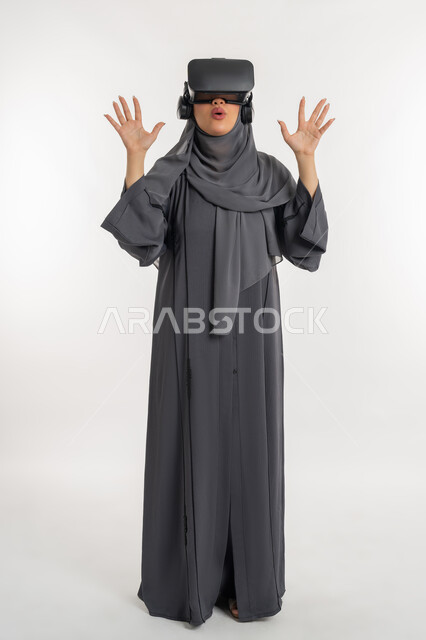 Using modern technology and techniques, simulating the imaginary world, a young Saudi woman raising her hands in amazement and surprise gestures, a portrait of a veiled Emirati Gulf Arab woman wearing an abaya and virtual reality glasses, interaction with a 3D video, full body length, white background