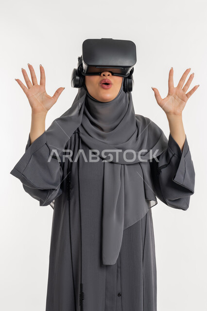 رفع اليدين بايماءات الدهشة والاستغراب, شابة سعودية تشاهد الأفلام عبر نظارات في آر، بورتريه لإمرأة عربية خليجية إماراتية محجبة ترتدي عباءة وتضع نظارة الواقع الافتراضي، التفاعل مع فيديو ثلاثي الأبعاد، استخدام التكنولوجيا الحديثة، محاكاة العالم الخيالي، خلفيه بيضاء