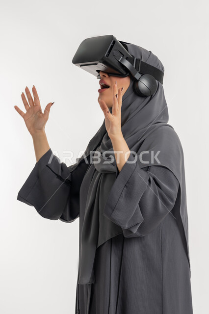 محاكاة العالم الخيالي، شابة سعودية ترفع يديها بايماءات الدهشة والاستغراب, مشاهدة الأفلام عبر نظارات في آر، بورتريه لإمرأة عربية خليجية إماراتية محجبة ترتدي العباءة تضع نظارة الواقع الافتراضي، التفاعل مع فيديو ثلاثي الأبعاد، استخدام التكنولوجيا الحديثة، خلفيه بيضاء
