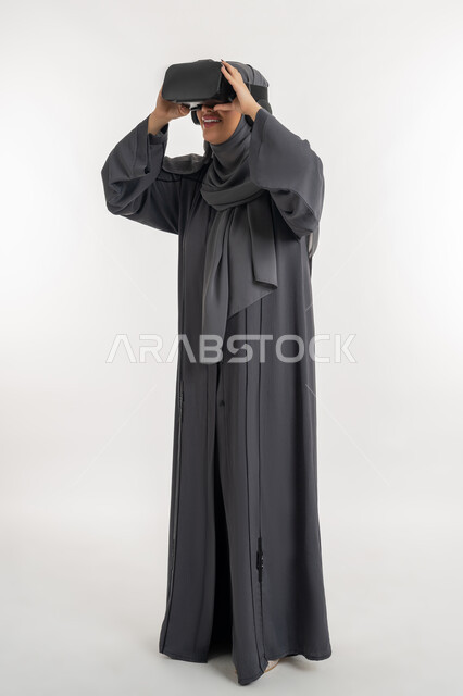 التسلية بالمشاهدة عبر نظارة الواقع الافتراضي،  بورتريه لإمرأة عربية خليجية إماراتية محجبة ترتدي العباءة وتنظرعبر نظارات ثلاثية الأبعاد، الاندماج ومحاكاة العالم الخيالي ، شابة سعودية بايماءات السعادة والسرور، التطور التقني والتكنولوجي, الطول الكامل للجسم، خلفيه يضاء