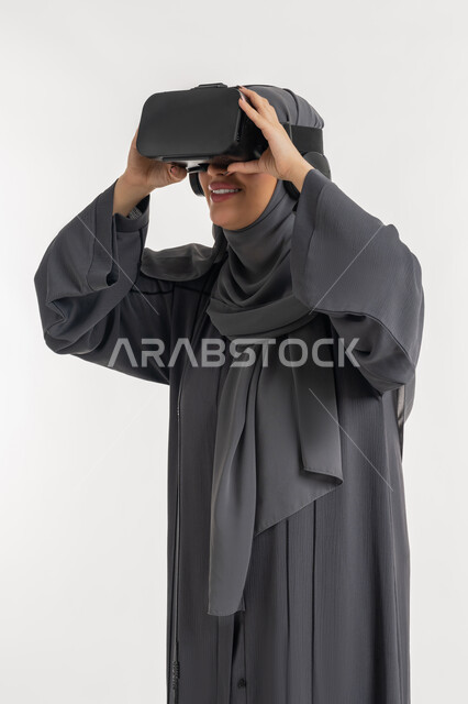 التطور التقني والتكنولوجي, المتعة و التسلية بالمشاهدة عبر نظارة الواقع الافتراضي،  بورتريه لإمرأة عربية خليجية إماراتية محجبة ترتدي العباءة وتنظر من خلال نظارات ثلاثية الأبعاد، الاندماج ومحاكاة العالم الخيالي ، شابة سعودية بايماءات السعادة والسرور، خلفيه يضاء