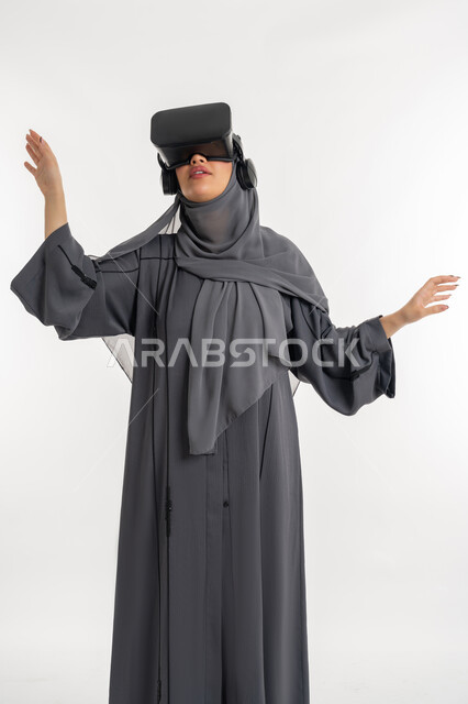 Integration and interaction with virtual reality, fun and entertainment by simulating the imaginary world, using advanced devices and technology, portrait of a veiled Emirati Gulf Arab woman wearing an abaya and 3D glasses, a young Saudi woman raising her hands and trying to touch something, white background