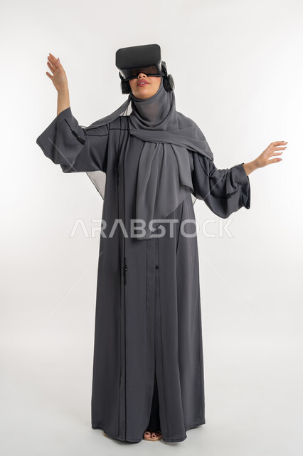 المتعة بمحاكاة العالم الخيالي, استخدام الأجهزة الحديثة والمتطورة, بورتريه لامرأة عربية خليجية إماراتية محجبة ترتدي عباءة وتضع نظارة ثلاثية الأبعاد، شابة سعودية ترفع يديها وتحاول لمس شيء ما، الاندماج والتفاعل مع الواقع الافتراضي، الطول الكامل للجسم، خلفية بيضاء