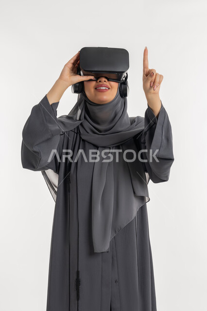 محاولة النقر على شيء ما, المتعة بالمشاهدة عبر نظارة الواقع الافتراضي, بورتريه لإمرأة عربية خليجية إماراتية محجبة ترتدي العباءة تضع نظارات ثلاثية الأبعاد, الاندماج ومحاكاة العالم الخيالي, شابة سعودية ترفع اصبع السبابة وتشير الى مكان معين، خلفية بيضاء