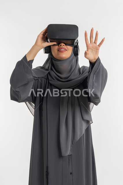 Simulating the imaginary world through VR glasses, portrait of a veiled Gulf Arab Emirati woman wearing an abaya, putting on 3D glasses and moving and interacting with them, a young Saudi woman raising her hands with gestures of integration, watching movies using virtual reality technologies, white background