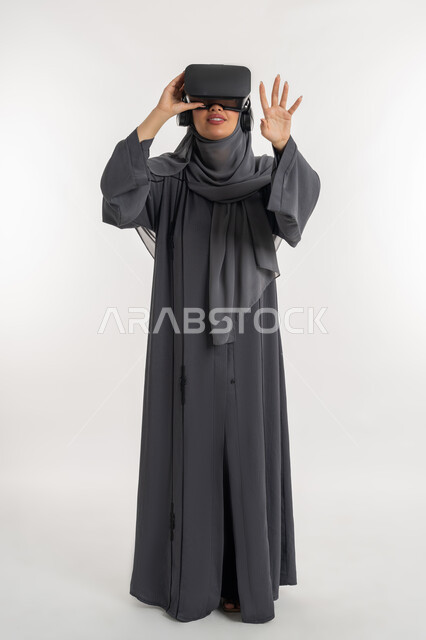 استخدام تقنيات الواقع الافتراضي, محاكاة العالم الخيالي عبر نظارات الفي آر, شابة سعودية ترفع يديها بإيماءات الاندماج والاستمتاع، بورتريه لإمرأة عربية خليجية إماراتية محجبة ترتدي العباءة تضع نظارة ثلاثية الأبعاد وتقوم بالتفاعل معها، الطول الكامل للجسم، خلفية بيضاء