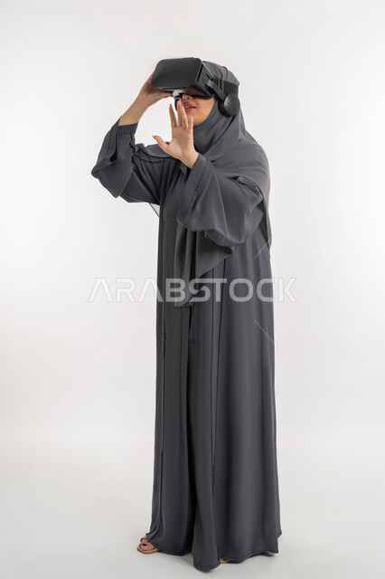 رفع اليدين بإيماءات الاندماج والاستمتاع، محاكاة العالم الخيالي, بورتريه لإمرأة عربية خليجية إماراتية محجبة ترتدي العباءة تضع نظارة ثلاثية الأبعاد وتقوم بالتحرك والتفاعل معها، شابة سعودية تشاهد الأفلام باستخدام تقنيات الواقع الافتراضي, الطول الكامل للجسم، خلفية بيضاء