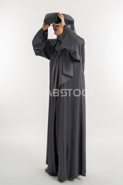 التسلية بالمشاهدة عبر نظارة الواقع الافتراضي،  بورتريه لإمرأة عربية خليجية إماراتية محجبة ترتدي العباءة وتنظر من خلال نظارات ثلاثية الأبعاد، الاندماج ومحاكاة العالم الخيالي ، شابة سعودية بايماءات السعادة والسرور، التطور التقني والتكنولوجي, الطول الكامل للجسم، خلفيه بيضاء