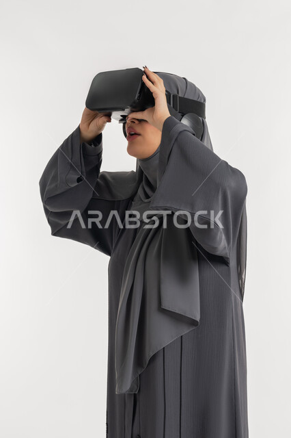 التطور التقني والتكنولوجي, المتعة و التسلية بالمشاهدة عبر نظارة الواقع الافتراضي،  بورتريه لإمرأة عربية خليجية إماراتية محجبة ترتدي العباءة وتنظر من خلال نظارات ثلاثية الأبعاد، شابة سعودية بايماءات السعادة والسرور، الاندماج ومحاكاة العالم الخيالي ، خلفيه بيضاء