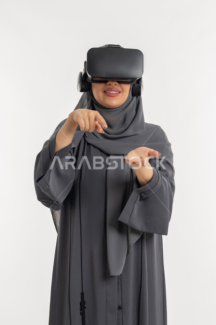 المتعة والتسلية بالمشاهدة عبر نظارة الواقع الافتراضي, بورتريه لإمرأة عربية خليجية إماراتية محجبة ترتدي العباءة تضع نظارات ثلاثية الأبعاد, شابة سعودية تشير باصبع السبابة الى كف اليد, ايماءات السعادة والسرور، الاندماج ومحاكاة العالم الخيالي، خلفية بيضاء