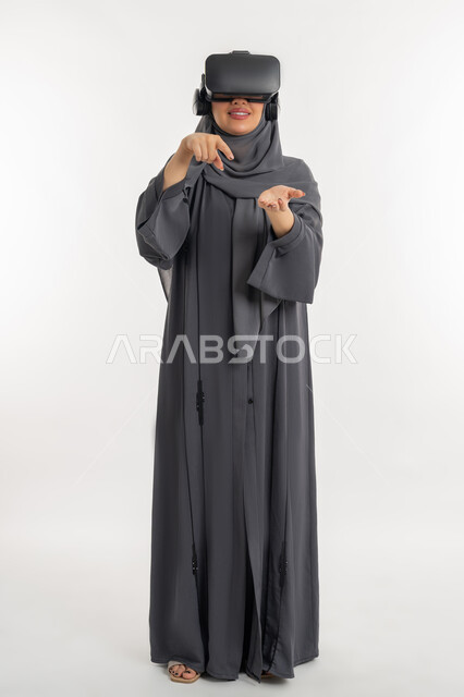 Index finger pointing to palm, merging with imaginary world, enjoying watching through virtual reality glasses, portrait of a veiled Emirati Gulf Arab woman wearing an abaya and 3D glasses, a young Saudi woman with gestures of happiness and pleasure, full body length, white background