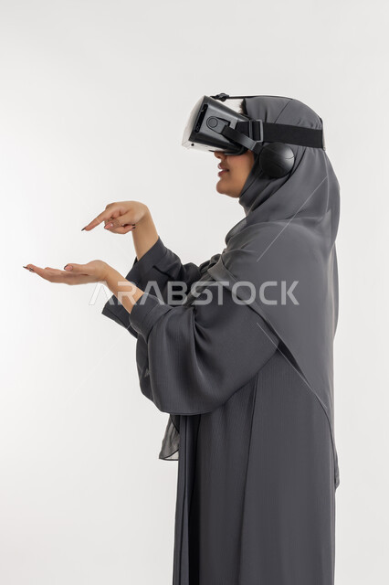 الاندماج ومحاكاة العالم الخيالي، المتعة والتسلية بالمشاهدة عبر نظارة الواقع الافتراضي, بورتريه من الجانب لإمرأة عربية خليجية إماراتية محجبة ترتدي العباءة تضع نظارات ثلاثية الأبعاد, شابة سعودية تشير باصبع السبابة الى كف اليد, ايماءات السعادة والسرور، خلفية بيضاء