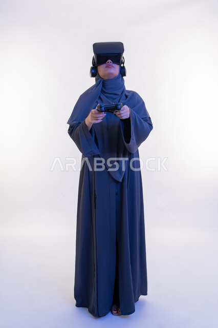 Using modern devices and technologies, expressions of victory and triumph, merging entertainment with the imaginary world, portrait of a veiled Emirati Gulf Arab woman wearing an abaya and virtual reality glasses holding a joystick, a young Saudi woman playing recreational video games, full body length, white background
