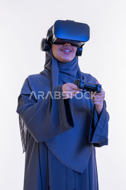 قضاء وقت ممتع بالألعاب الالكترونية الترفيهية، الإندماج والتفاعل مع الجيم، دمج التسلية مع العالم الخيالي، بورتريه لإمرأة عربية خليجية إماراتية محجبة ترتدي العباءة ونظارة الواقع الإفتراضي تمسك عصا التحكم، شابة سعودية تستخدم الأجهزة والتقنيات الحديثة، خلفية بيضاء