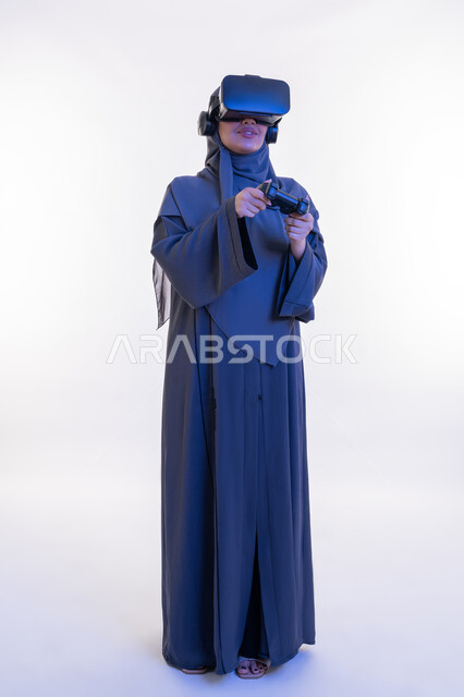 استخدام الأجهزة والتقنيات الحديثة، تعابير الانتصار والفوز، دمج التسلية مع العالم الخيالي، بورتريه لإمرأة عربية خليجية إماراتية محجبة ترتدي عباءة ونظارة الواقع الإفتراضي تمسك عصا التحكم، شابة سعودية تلعب بألعاب الفيديو الترفيهية، الطول الكامل للجسم، خلفية بيضاء