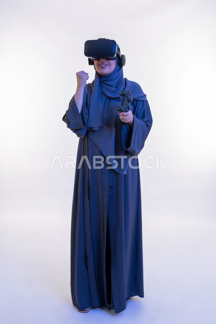 أجهزة تكنولوجية وتقنيات حديثة، تعابير الانتصار والفوز، دمج المتعة مع العالم الخيالي، بورتريه لإمرأة عربية خليجية إماراتية محجبة ترتدي العباءة ونظارة الواقع الإفتراضي تمسك بيدها عصا التحكم، شابة سعودية تلعب بألعاب الفيديو الترفيهية، الطول الكامل للجسم، خلفية بيضاء