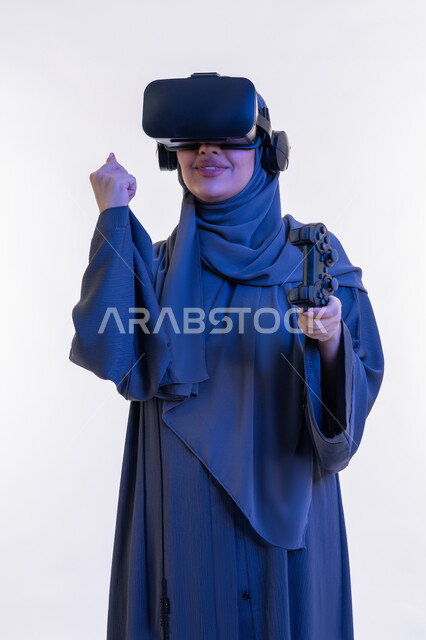 دمج المتعة والتسلية مع العالم الخيالي، بورتريه لإمرأة عربية خليجية إماراتية محجبة ترتدي العباءة ونظارة الواقع الإفتراضي تمسك بيدها عصا التحكم، شابة سعودية تلعب بألعاب الفيديو الترفيهية، أجهزة تكنولوجية وتقنيات حديثة، تعابير الانتصار والفوز، خلفية بيضاء