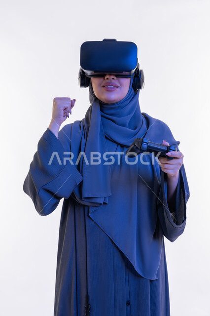 تعابير الانتصار والفوز، دمج المتعة والتسلية مع العالم الخيالي، بورتريه لإمرأة عربية خليجية إماراتية محجبة ترتدي العباءة ونظارة الواقع الإفتراضي تمسك بيدها عصا التحكم، شابة سعودية تلعب بألعاب الفيديو الترفيهية، أجهزة تكنولوجية وتقنيات حديثة، خلفية بيضاء