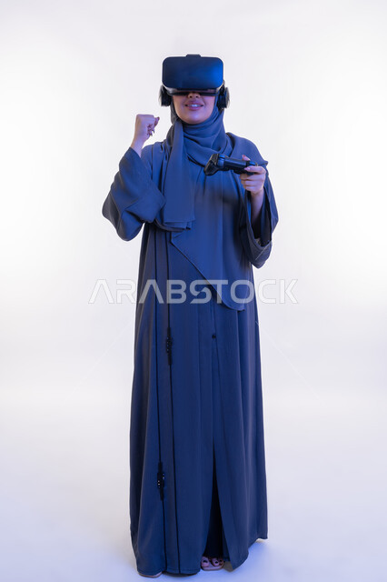 لعب ألعاب الفيديو الترفيهية، أجهزة تكنولوجية وتقنيات حديثة، دمج المتعة مع العالم الخيالي، بورتريه لإمرأة عربية خليجية إماراتية محجبة ترتدي العباءة ونظارة الواقع الإفتراضي تمسك بيدها عصا التحكم، شابة سعودية بتعابير الانتصار والفوز، الطول الكامل للجسم، خلفية بيضاء
