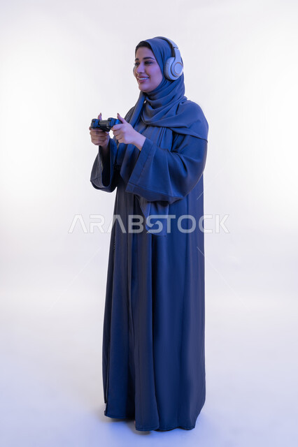 إيماءات الاستمتاع والاندماج والتفاعل, قضاء إجازة صيفية ممتعة، تقنيات الكترونية حديثة, بورتريه لإمرأة عربية خليجية إماراتية محجبة ترتدي العباءة تضع سماعة الهيدفون وتحمل بيدها عصا التحكم، شابة سعودية تلعب بألعاب الفيديو الترفيهية، الطول الكامل للجسم، خلفية بيضاء