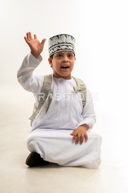 القاء التحية والسلام، مفهوم الاناقة والاهتمام بالمظهر الخارجي, العودة الى المدرسة بحماس، بورتريه لولد عربي خليجي عماني يرتدي الدشداشة و الكمه يحمل حقيبة الظهر المدرسية يرفع يده بايماءات الترحيب والاستقبال، خلفية بيضاء