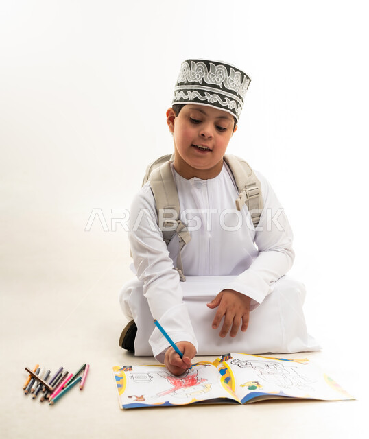 Developing artistic skills for people with disabilities, using wooden crayons, portrait of an Arab Gulf Omani boy with Down syndrome wearing a dishdasha and a cloak, carrying a backpack, learning to draw and color with focus and precision, supporting the Omani community for people with special needs, white background