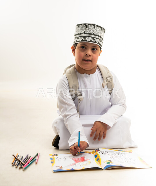 Supporting the Omani community for people with special needs, developing the artistic skills of people with disabilities, using wooden crayons, portrait of an Arab Gulf Omani boy with Down syndrome wearing a dishdasha and a cloak, carrying a backpack, learning to draw and color with focus and precision, white background