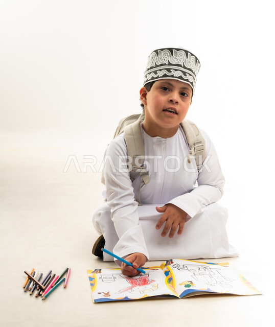 استخدام أقلام التلوين الخشبية, تنمية المهارة الفنية لذوي الهمم, بورتريه لصبي عربي خليجي عماني مصاب بمتلازمة داون يرتدي الدشداشة والكمة يحمل حقيبة الظهر يقوم بتعلم الرسم والتلوين بتركيز ودقة، دعم المجتمع العماني لذوي الاحتياجات الخاصة, خلفية بيضاء