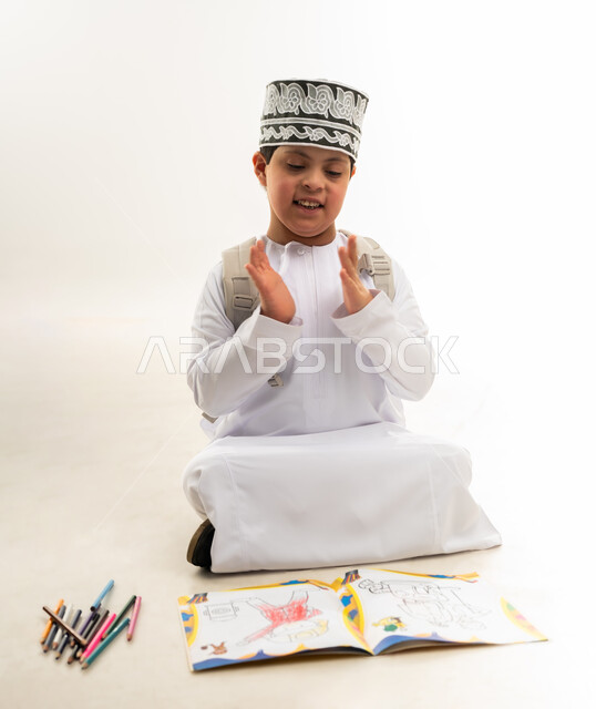 تنمية المهارة الفنية لذوي الهمم, استخدام أقلام التلوين الخشبية, الاستمتاع بالرسم والتلوين، بورتريه لصبي عربي خليجي عماني مصاب بمتلازمة داون يرتدي الدشداشة والكمة يحمل حقيبة الظهر يرفع يديه بايماءات التصفيق، خلفية بيضاء