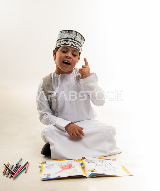 الاستمتاع بالرسم والتلوين، تنمية المهارة الفنية لذوي الهمم, استخدام أقلام التلوين الخشبية, بورتريه لصبي عربي خليجي عماني مصاب بمتلازمة داون يرتدي الدشداشة والكمة يحمل حقيبة الظهر يرفع اصبعه بايماءات الرقم واحد، خلفية بيضاء
