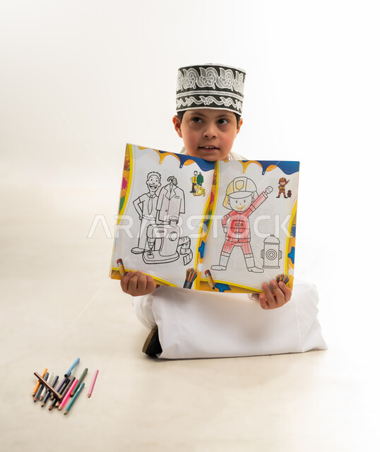 دعم المجتمع العماني لذوي الاحتياجات الخاصة, تنمية المهارة الفنية لذوي الهمم, استخدام أقلام التلوين الخشبية, بورتريه لصبي عربي خليجي عماني مصاب بمتلازمة داون يرتدي الدشداشة والكمة يحمل حقيبة الظهر يقوم بعرص لوحته، خلفية بيضاء