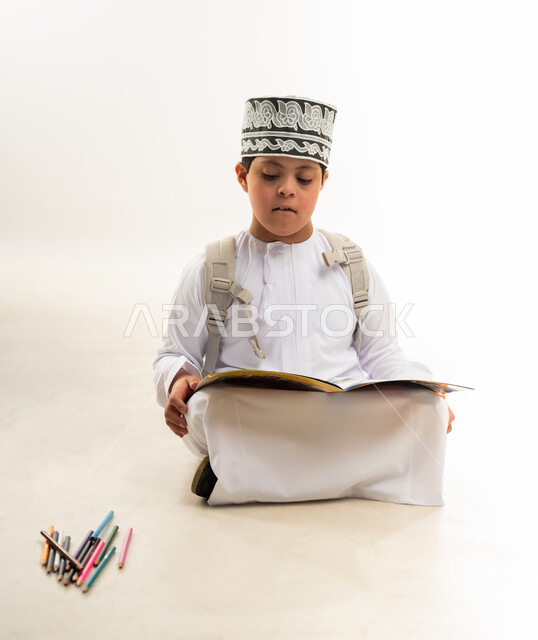 استخدام أقلام التلوين الخشبية, تنمية المهارة الفنية لذوي الهمم, بورتريه لصبي عربي خليجي عماني مصاب بمتلازمة داون يرتدي الدشداشة والكمة يحمل حقيبة الظهر يقوم بتعلم الرسم والتلوين بتركيز ودقة، دعم المجتمع العماني لذوي الاحتياجات الخاصة, خلفية بيضاء