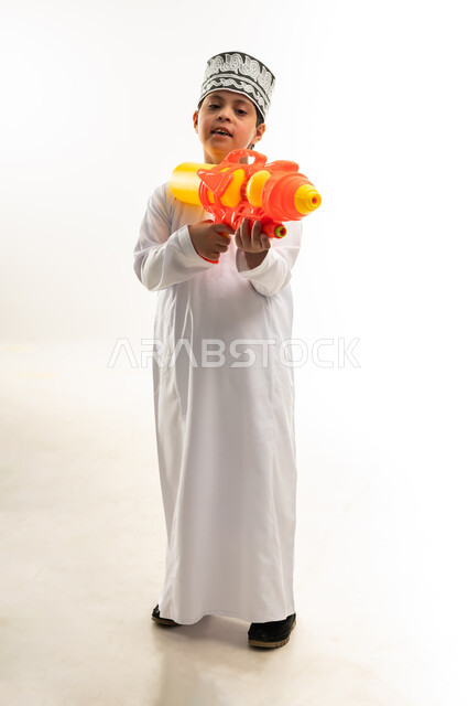 قضاء اوقات ممتعه بالألعاب المائية في فصل الصيف، مفهوم الترفيه عن النفس والتسلية، اسلوب حياة ذوي الهمم، بورتريه لشاب عربي خليجي عماني مصاب بمتلازمة داون يرتدي الدشداشة والكمة يحمل بيده مسدس مائي بلاستيكي يقوم باللعب بإيماءات السرور، صورة لكامل الجسم، خلفية بيضاء