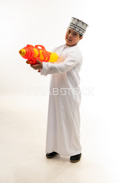 Lifestyle of people with disabilities, having fun with water games in the summer, the concept of entertainment and amusement, portrait of a young Gulf Arab Omani man with Down syndrome wearing a dishdasha and a kimono, holding a plastic water gun in his hand, playing with gestures of pleasure, full body image, white background