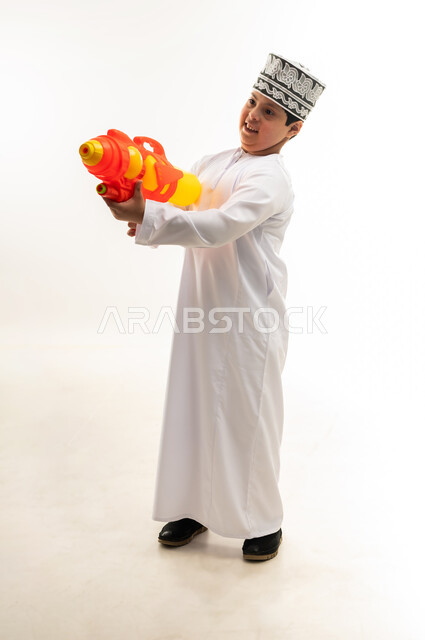 قضاء اوقات ممتعه بالألعاب المائية في فصل الصيف، مفهوم الترفيه عن النفس والتسلية، اسلوب حياة ذوي الهمم، بورتريه لشاب عربي خليجي عماني مصاب بمتلازمة داون يرتدي الدشداشة والكمة يحمل بيده مسدس مائي بلاستيكي يقوم باللعب بإيماءات السرور، صورة لكامل الجسم، خلفية بيضاء