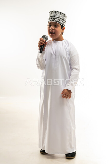 تسجيل مقطع صوتي غنائي، الاستمتاع بممارسة المواهب الفنية، قضاء وقت ممتع بأداء العروض الصوتية، بورتريه لصبي عربي خليجي عماني مصاب بمتلازمة داون يرتدي الدشداشة والكمة يحمل المايكروفون يقوم بالغناء بايماءات الاندماج، صورة لكامل الجسم، خلفية بيضاء