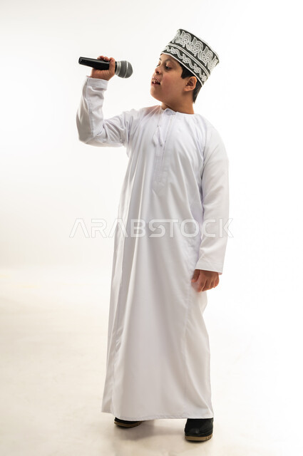 الاستمتاع بممارسة المواهب الفنية، قضاء وقت ممتع بأداء العروض الصوتية، تسجيل مقطع صوتي غنائي، بورتريه لصبي عربي خليجي عماني مصاب بمتلازمة داون يرتدي الدشداشة والكمة يحمل المايكروفون يقوم بالغناء بايماءات الاندماج، صورة لكامل الجسم، خلفية بيضاء