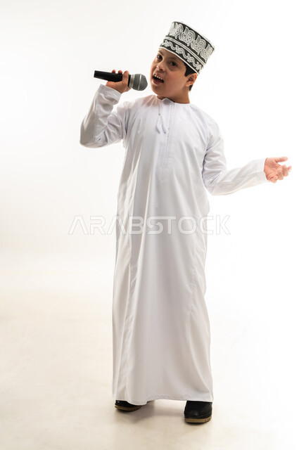 قضاء وقت ممتع بأداء العروض الصوتية، الاستمتاع بممارسة المواهب الفنية، تسجيل مقطع صوتي غنائي، بورتريه لصبي عربي خليجي عماني مصاب بمتلازمة داون يرتدي الدشداشة والكمة يحمل المايكروفون يرفع يده بايماءات الاندماج مع الغناء، صورة لكامل الجسم، خلفية بيضاء