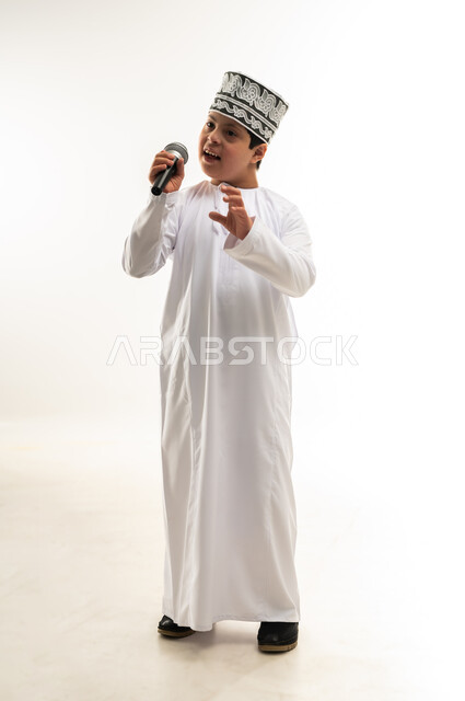 الاستمتاع بممارسة المواهب الفنية، قضاء وقت ممتع بأداء العروض الصوتية، تسجيل مقطع صوتي غنائي، بورتريه لصبي عربي خليجي عماني مصاب بمتلازمة داون يرتدي الدشداشة والكمة يحمل المايكروفون يرفع يده بايماءات الاندماج مع الغناء، صورة لكامل الجسم، خلفية بيضاء