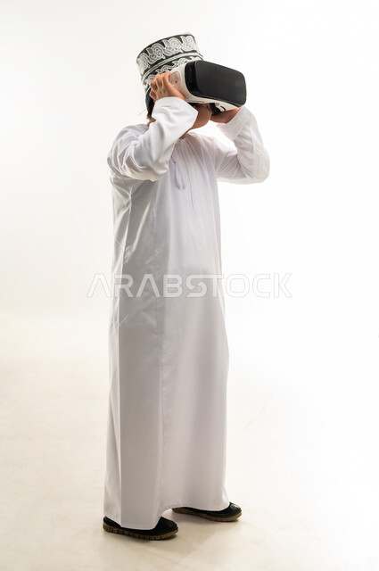تعابير الاندماج والتركيز، بورتريه لولد عربي خليجي عمانبي مصاب بمتلازمة داون يرتدي الدشداشة والكمة يقوم بمشاهدة الأفلام عبر نظارات الواقع الافتراضي ثلاثية الأبعاد، محاكاة العالم الخيالي والتفاعل معه، صورة لكامل الجسم، خلفيه بيضاء