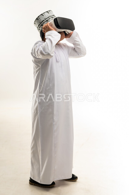 ايماءات الاندماج والتركيز، بورتريه لولد عربي خليجي عمانبي مصاب بمتلازمة داون يرتدي الدشداشة والكمة يقوم بمشاهدة الأفلام عبر نظارات الواقع الافتراضي ثلاثية الأبعاد، محاكاة العالم الخيالي والتفاعل معه، صورة لكامل الجسم، خلفيه بيضاء