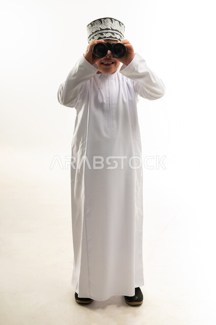 حب المغامرة والاستكشاف، رؤية الاشياء البعيدة، اسلوب حياة أصحاب الهمم، شعور المعاقين بالإنتماء والتقدير، بورتريه لصبي عربي خليجي عماني يرتدي الدشداشة والكمة مصاب بمتلازمة داون ينظر من خلال المنظار ثنائي العينين، صورة لكامل الجسم، خلفية بيضاء