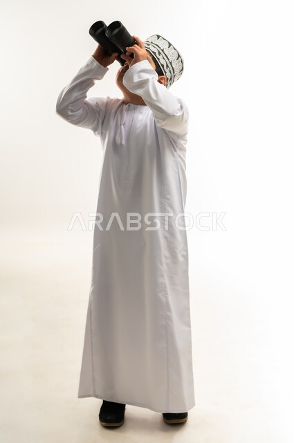 رؤية الاشياء البعيدة، حب المغامرة والاستكشاف، اسلوب حياة أصحاب الهمم، شعور المعاقين بالإنتماء والتقدير، بورتريه لصبي عربي خليجي عماني يرتدي الدشداشة والكمة مصاب بمتلازمة داون ينظر من خلال المنظار ثنائي العينين، صورة لكامل الجسم، خلفية بيضاء