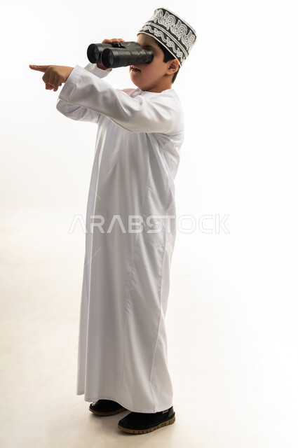 شعور المعاقين بالإنتماء والتقدير، رؤية الاشياء البعيدة، حب المغامرة والاستكشاف، بورتريه من الجانب لصبي عربي خليجي عماني يرتدي الدشداشة والكمة مصاب بمتلازمة داون ينظر من خلال المنظار ثنائي العينين يشير باصبع السبابة لمكان ما، صورة لكامل الجسم، خلفية بيضاء