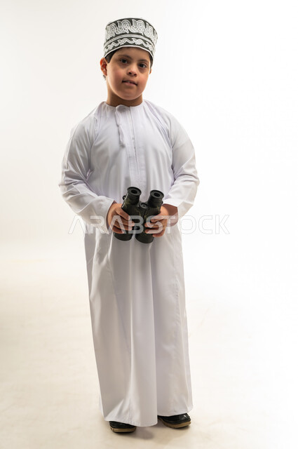 اسلوب حياة أصحاب الهمم، رؤية الاشياء البعيدة، حب المغامرة والاستكشاف، شعور المعاقين بالإنتماء والتقدير، بورتريه لصبي عربي خليجي عماني يرتدي الدشداشة والكمة مصاب بمتلازمة داون يحمل بيده المنظار ثنائي العينين، صورة لكامل الجسم، خلفية بيضاء