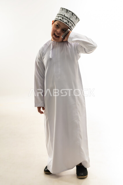 اجراء المكالمات الهاتفية، ايماءات السرور والسعادة، تعليم المعاقين على استخدام الأجهزة التقنية المتطورة، بورتريه لصبي عربي خليجي عماني مبتسم مصاب بمتلازمة داون يرتدي الدشداشة والكمة يحمل بيده الهاتف المحمول، صورة لكامل الجسم، خلفية بيضاء
